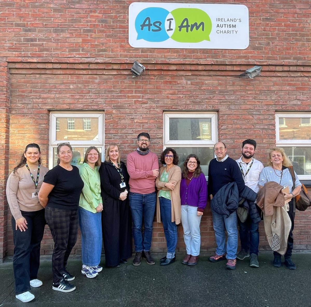 Skupina od deset odraslih ljudi stoji nasmiješeno ispred zidane zgrade. Iznad njih je natpis 'AsIAm – Ireland's Autism Charity'. Osobe stoje u nizu, opuštene su i izgledaju kao da sudjeluju u zajedničkom posjetu ili programu.