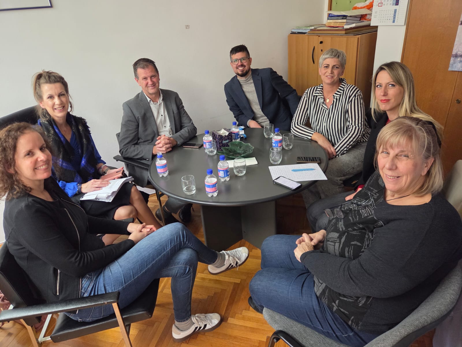 Skupina od sedam osoba sjedi oko okruglog stola u uredu, nasmijani gledaju u kameru, na stolu su bočice vode, čaše, papiri i mobiteli, što sugerira radni sastanak u opuštenoj atmosferi.