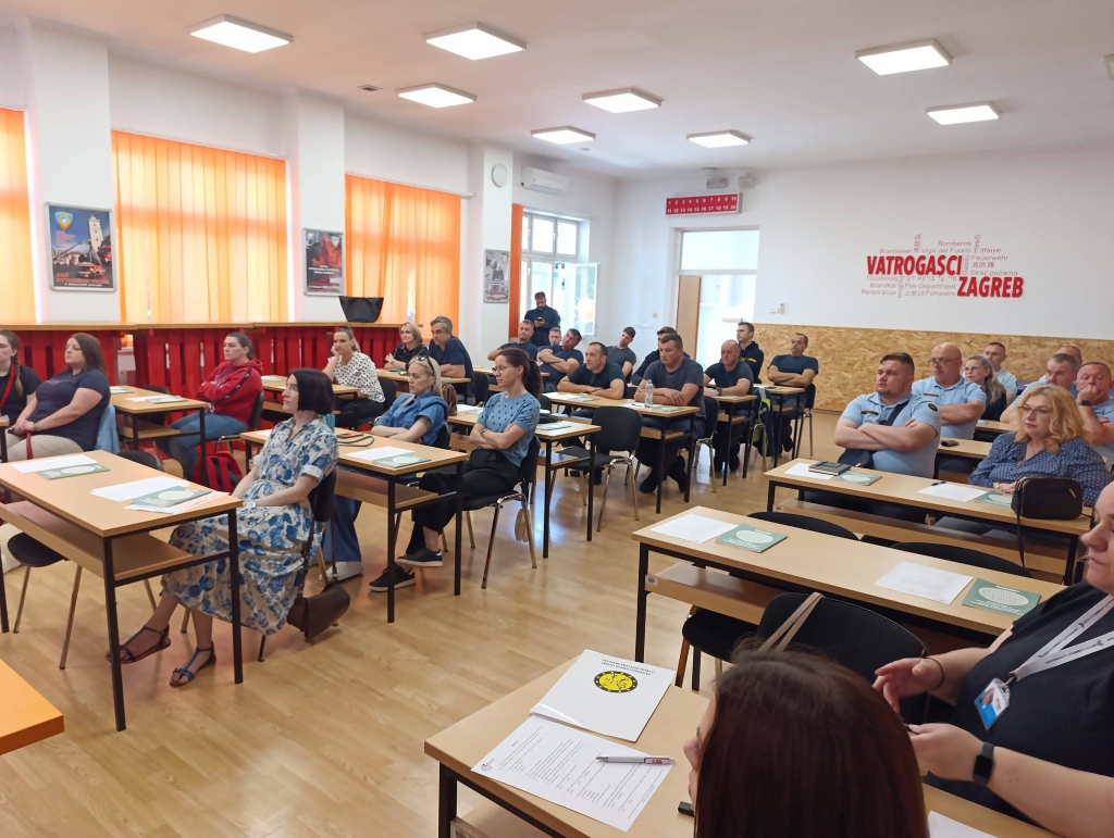 Sudionici edukacije sjede u učionici za stolovima i prate predavanje. Na zidu je vidljiv natpis „Vatrogasci Zagreb”, a na stolovima su raspoređeni edukativni materijali.