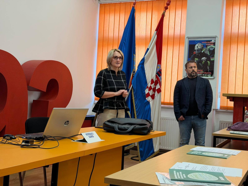Dvije osobe, jedna žena i jedan muškarac, stoje na početku edukacije u učionici i obraćaju se sudionicima. U prostoru su vidljivi stolovi s materijalima, prijenosno računalo te zastave Grada Zagreba i Republike Hrvatske.