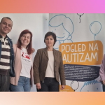 Četvero ljudi stoji u zatvorenom prostoru pokraj plakata na kojem piše „Pogled na autizam“. Plakat prikazuje ilustrirane balone i crtež nasmijanog djeteta. Skupina se smiješi i pozira za fotografiju.