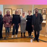 Šest osoba stoji nasmiješeno u muzeju ispred zida na kojem vise portreti i uokvireni dokumenti. Četiri žene i jedan muškarac stoje u prvom planu, dok se iza njih vidi drveni pod i izložbeni prostor s toplim osvjetljenjem. Pozadina slike ima blagi prijelaz boja od narančaste do ružičaste.