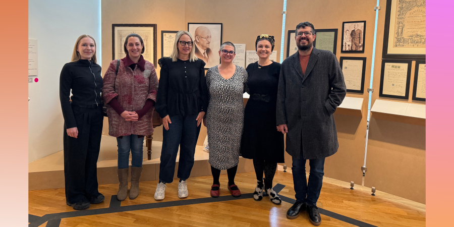 Šest osoba stoji nasmiješeno u muzeju ispred zida na kojem vise portreti i uokvireni dokumenti. Četiri žene i jedan muškarac stoje u prvom planu, dok se iza njih vidi drveni pod i izložbeni prostor s toplim osvjetljenjem. Pozadina slike ima blagi prijelaz boja od narančaste do ružičaste.