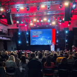 Velika konferencijska dvorana ispunjena sjedećim sudionicima okrenutima prema jako osvijetljenoj pozornici, na kojoj izlagač stoji za govornicom ispred velikog ekrana s naslovom “From Vision to Change Towards 2030” u modernom crveno-plavom dizajnu.