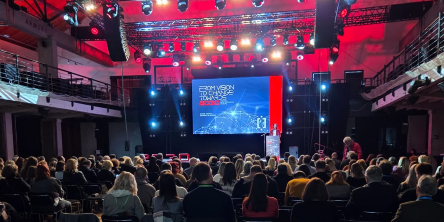 Velika konferencijska dvorana ispunjena sjedećim sudionicima okrenutima prema jako osvijetljenoj pozornici, na kojoj izlagač stoji za govornicom ispred velikog ekrana s naslovom “From Vision to Change Towards 2030” u modernom crveno-plavom dizajnu.