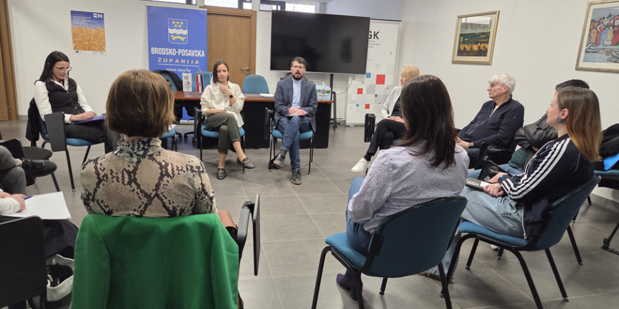 Grupa od desetak ljudi sjedi u krugu na plavim stolicama u uredskom prostoru i sudjeluje u raspravi. U sredini sjede muškarac u sakou i žena u svijetloj bluzi koji govore, dok ostali pažljivo slušaju. U pozadini se vidi banner s natpisom „Brodsko-posavska županija“, veliki ekran i zid s uokvirenim slikama.
