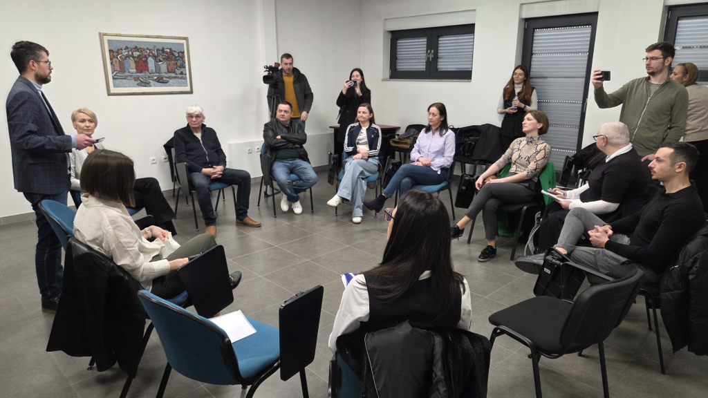 Muškarac u odijelu stoji i govori grupi od desetak ljudi koji sjede u krugu na stolicama u svijetloj prostoriji s bijelim zidovima. Sudionici ga pažljivo slušaju; neki drže bilježnice, a nekoliko osoba stoji sa strane i snima mobitelom i kamerom. Na zidu je uokvirena slika, a iza grupe su prozori sa spuštenim roletama. Atmosfera podsjeća na sastanak, radionicu ili konferenciju za medije.