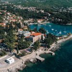 Fotografija iz zraka primorskog mjesta za sunčanog dana. Na rtu uz more stoji velika zgrada od crvenkaste opeke s narančastim krovom, okružena zelenilom. Na terasi ispred zgrade održava se okupljanje na otvorenom: ljudi sjede u redovima stolica. Uz stjenovitu obalu vodi kamena šetnica s malim betonskim kupalištem. Desno je zaljev s jedrilicama i brodicama usidrenima uz mol. Kuće s crvenim krovovima penju se uz šumovito brdo, a more je tirkizno i tamnoplavo.